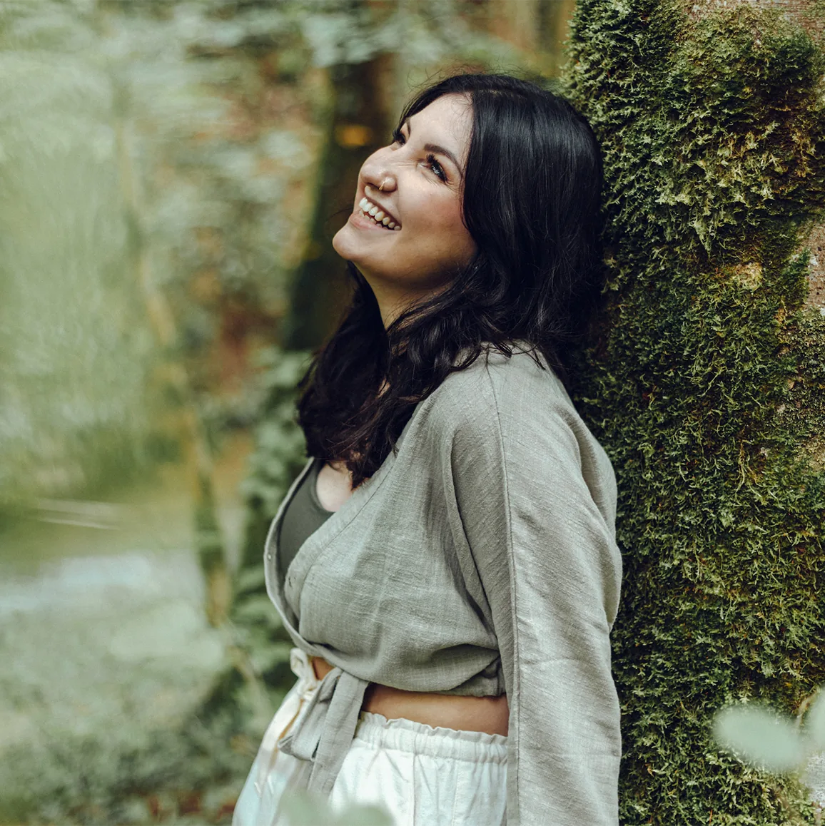 Yasemin, kinésiologue à Neuchâtel, souriante, appuyée contre un arbre couvert de mousse